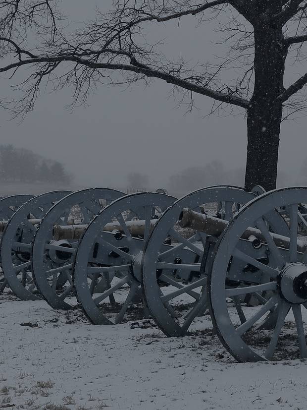A line of Revolutionary War cannons standing in snow at Artillery Park, Valley Forge, bare winter trees and low grey fog behind them.