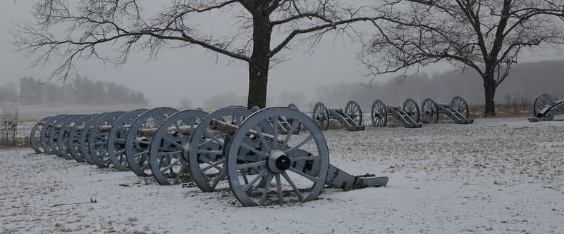 A line of Revolutionary War cannons under snow at Artillery Park, Valley Forge, with bare winter trees behind them.